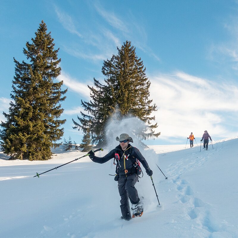 Zie een sneeuwschoenwandelaar bergafwaarts naar beneden lopen. Hij gaat op een hoog tempo dus je ziet sneeuw opspatten. In de achtergrond zie je twee naaldbomen.