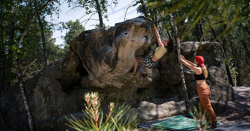 Twee vrouwen boulderen in Fontainebleau. Een vrouw hangt aan een steen en de andere vrouw spot de klimmende vrouw.