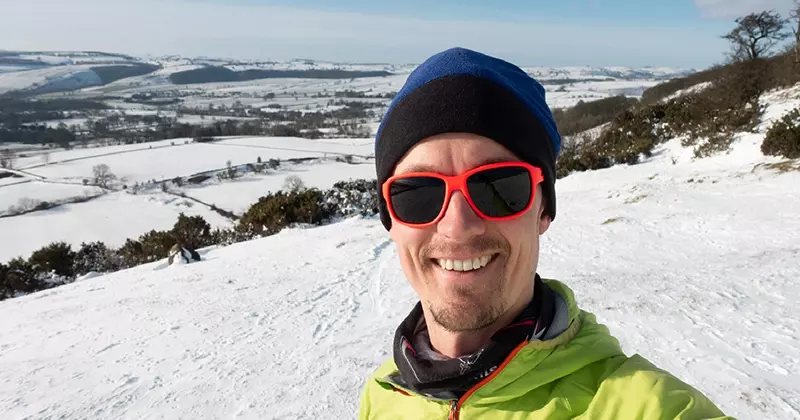Een man met een rode zonnebril en winterkleding lacht voor een selfie op een besneeuwde heuvel, met velden en heuvels op de achtergrond onder een blauwe lucht.