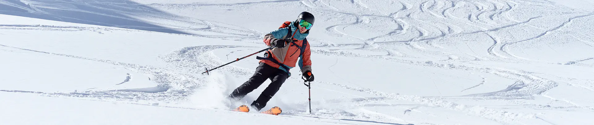 Een skiër in een blauwe jas, een rode broek en met een rugzak skiet onder een gedeeltelijk bewolkte hemel een steile, besneeuwde berghelling af, vlakbij een rotspartij.