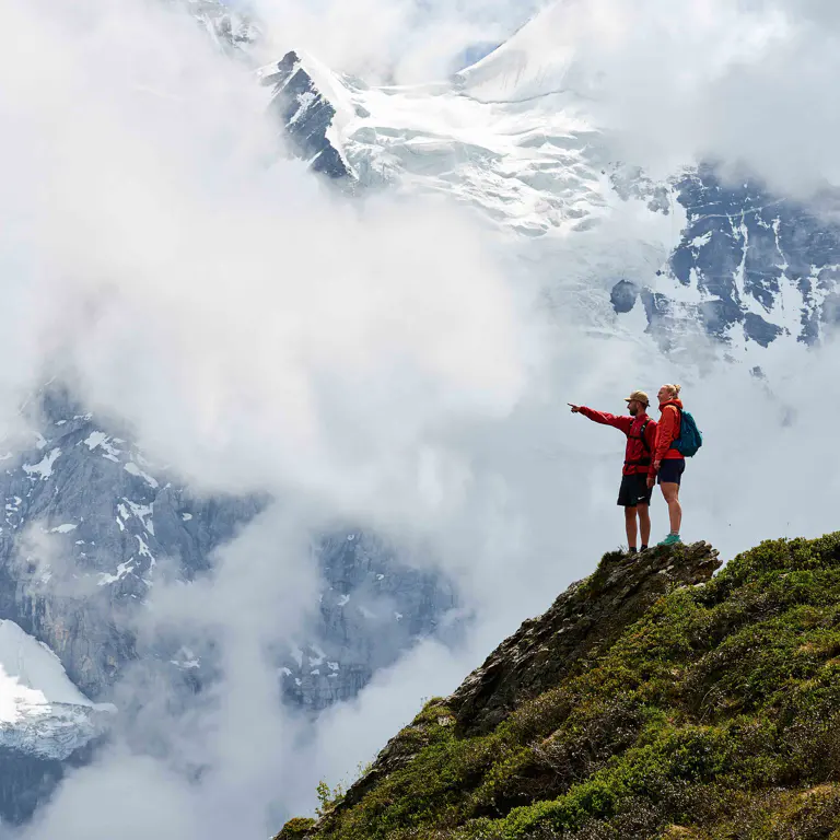Twee bergwandelaars staan op een bergkam en wijzen naar een met sneeuw bedekte bergtop. Twee bergwandelaars staan op een bergkam en wijzen naar een met sneeuw bedekte bergtop.
