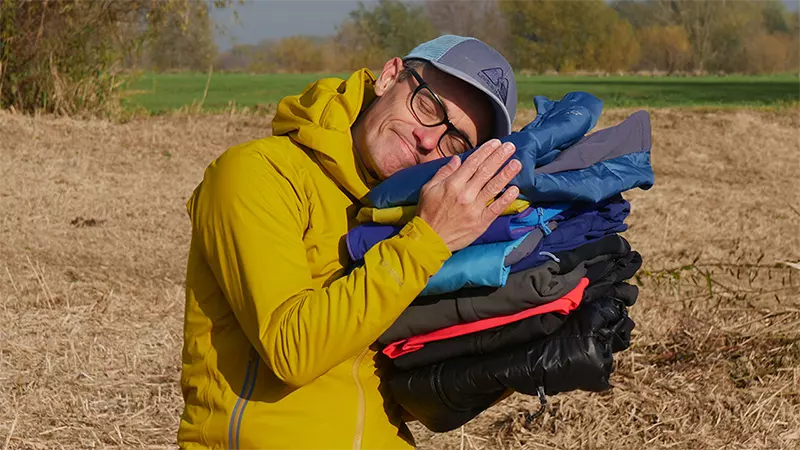 Een persoon in een geel jasje en een pet glimlacht terwijl hij buiten, in een groene, landelijke omgeving, een stapel kleurrijke, opgevouwen jasjes vasthoudt.