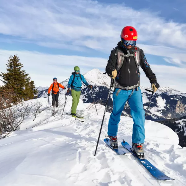 Drie mensen met helmen en winterkleding skiën bergopwaarts door de sneeuw op een berg, met bomen en bergtoppen op de achtergrond onder een gedeeltelijk bewolkte blauwe hemel.