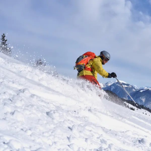 Een skiër in een geel jack en met een oranje rugzak glijdt door verse poedersneeuw op een besneeuwde berghelling, terwijl de sneeuw omhoog spuit, onder een blauwe hemel en met uitzicht op bergen in de verte.