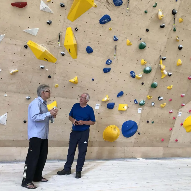 Twee oudere mannen staan te praten voor een klimwand binnenshuis die bedekt is met kleurrijke grepen. De ene draagt een licht shirt en houdt een waterfles vast, terwijl de andere een blauw T-shirt draagt. De klimwand is voorzien van gele, blauwe en zwarte grepen.