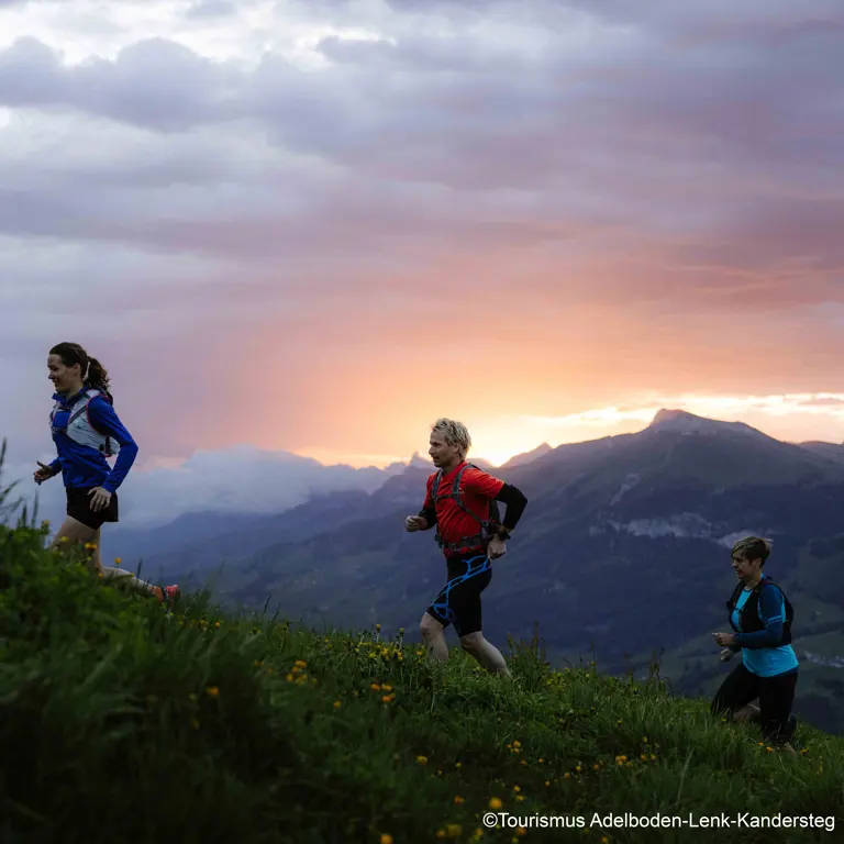 Drie mensen die bij zonsondergang bergopwaarts door een met gras begroeid, met bloemen bezaaid landschap trailrunnen, met indrukwekkende wolken en bergtoppen op de achtergrond.