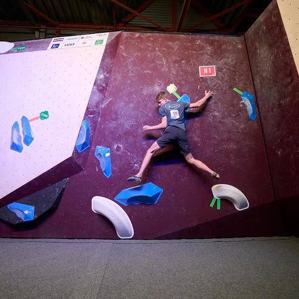Een jonge klimmer in een blauw shirt met nummer 11 beklimt tijdens een wedstrijd een kastanjebruine indoor-bouldermuur met blauwe en witte grepen. De scène is helder verlicht en boven de muur zijn diverse sponsorlogo’s te zien.