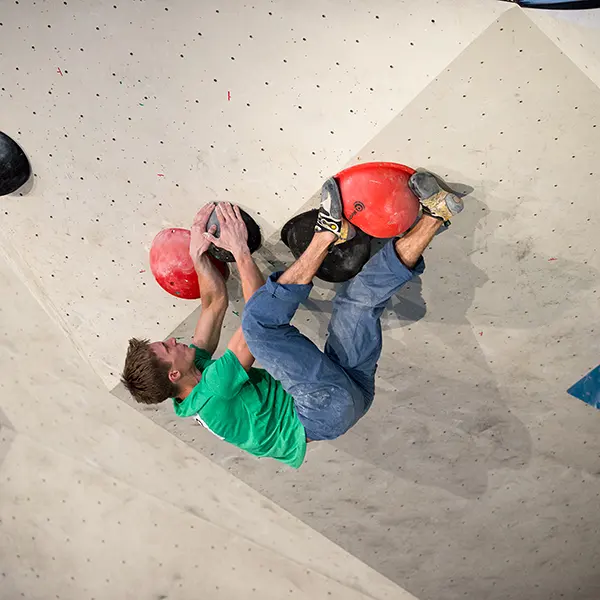 Een man in een groen shirt en een blauwe broek is aan het boulderen in een klimhal; hij houdt zich met zijn handen en voeten vast aan grote rode grepen op een klimwand.