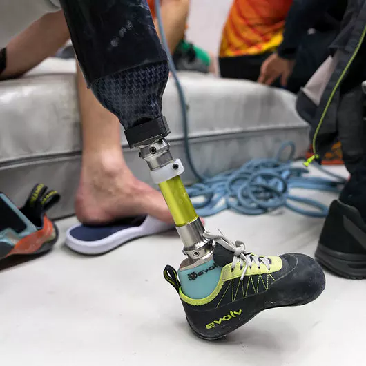 Een persoon met een beenprothese en klimschoenen aan staat naast klimuitrusting en andere mensen, klaar om te gaan klimmen in de klimhal.