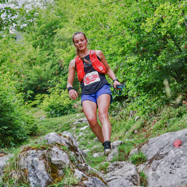 Een vrouw in een rood vest, een blauwe korte broek en met een startnummer rent tijdens een trailrace over een rotsachtig pad, omringd door groene bomen en begroeiing.