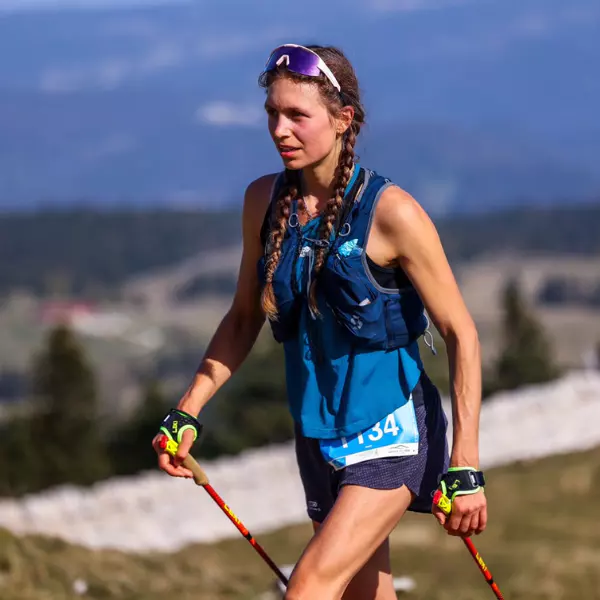 Een vrouwelijke trailrunner in sportkleding en met een zonnebril op gebruikt wandelstokken terwijl ze in de buitenlucht bergopwaarts loopt, met op de achtergrond een wazig berglandschap.