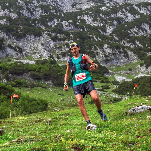 Een trailrunner met een startnummer, een vest en een zonnebril rent over een met gras begroeid, rotsachtig bergpad, met op de achtergrond steile, groene heuvels en oranje markeringen.