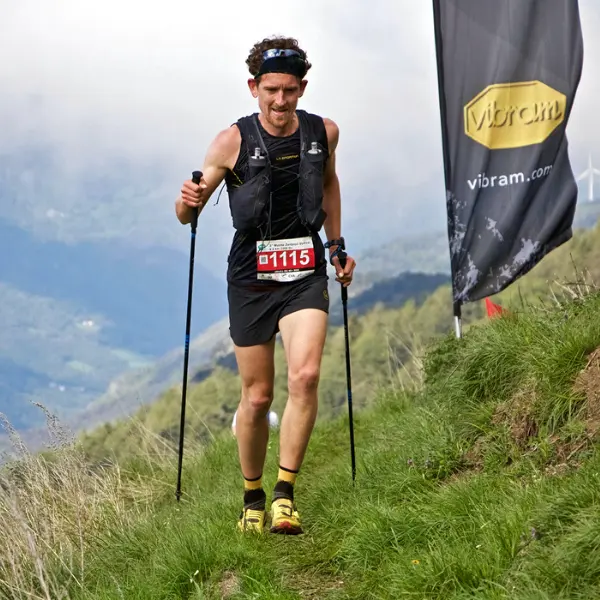 Een mannelijke trailrunner met wandelstokken, met startnummer 1115, een zwart vest en gele schoenen, rent bergopwaarts over een met gras begroeid bergpad vlakbij een Vibram-spandoek.