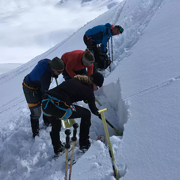 Vier mensen in winterkleding graven met schoppen en sondes in de diepe sneeuw op een helling; ze zijn mogelijk bezig met