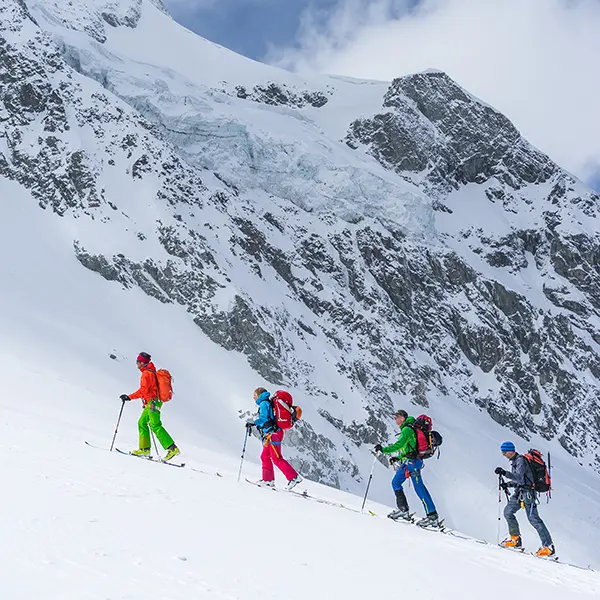 Vier skiërs in kleurrijke uitrusting klimmen omhoog over besneeuwde berghellingen, met rugzakken en skistokken in de hand, tegen een achtergrond van ruige, rotsachtige bergtoppen en glinsterende sneeuw.
