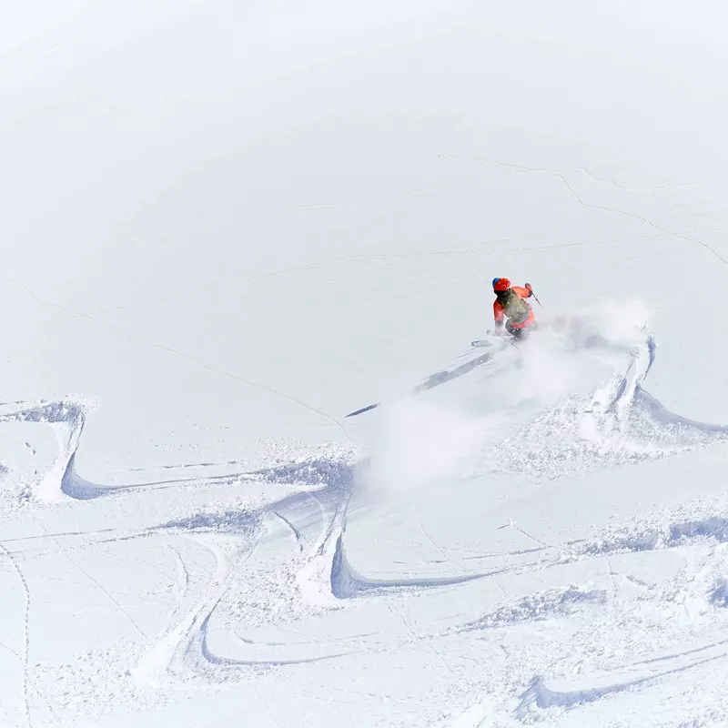 Een skiër in een rode jas en met een helm snijdt door de verse sneeuw en laat poedersporen achter op een besneeuwde helling onder een stralende hemel.