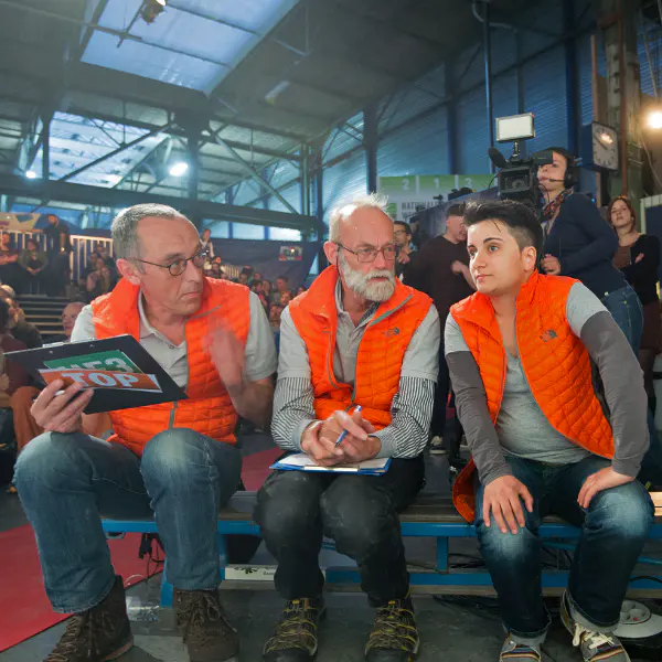 Drie mensen in oranje hesjes zitten binnen op een bankje; ze praten en houden klemborden vast. Op de achtergrond zijn een menigte, camera’s en een productieteam te zien, wat doet vermoeden dat het om een live-evenement of een tv-programma gaat.