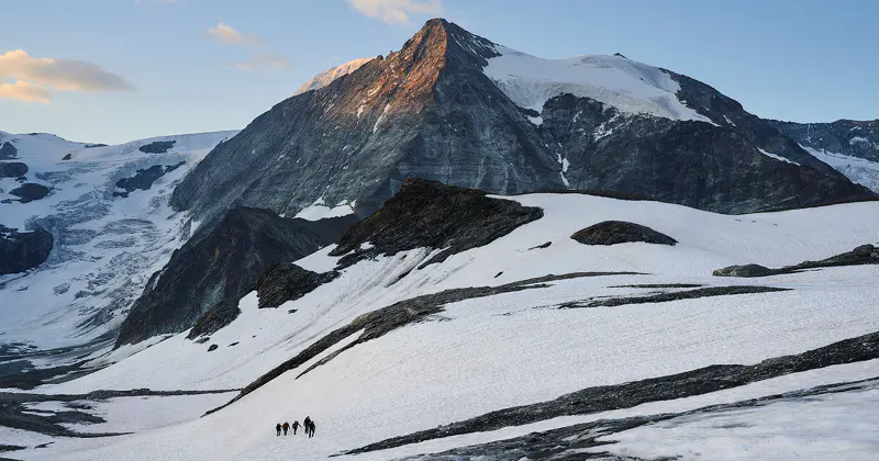 Vier wandelaars lopen over een besneeuwde berghelling onder een torenhoge, rotsachtige bergtop met stukken ijs, badend in het zachte licht van de vroege ochtend of avond.