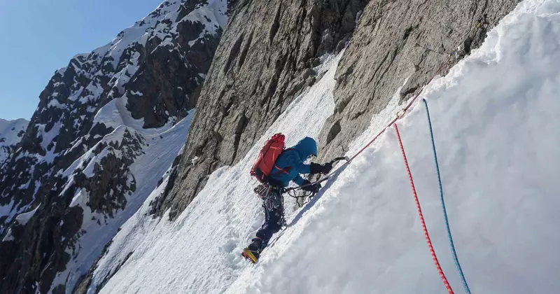 Een bergbeklimmer in winterkleding en met een rode rugzak beklimt een besneeuwde, steile berghelling met behulp van touwen en stijgijzers, met ruige, besneeuwde bergtoppen op de achtergrond.