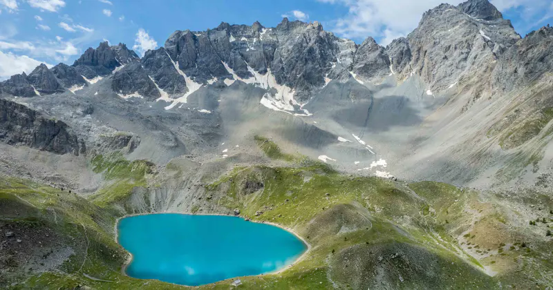 Een helder turkoois bergmeer wordt omringd door groene hellingen en rotsachtige, met sneeuw bestrooide bergtoppen onder een gedeeltelijk bewolkte blauwe hemel.