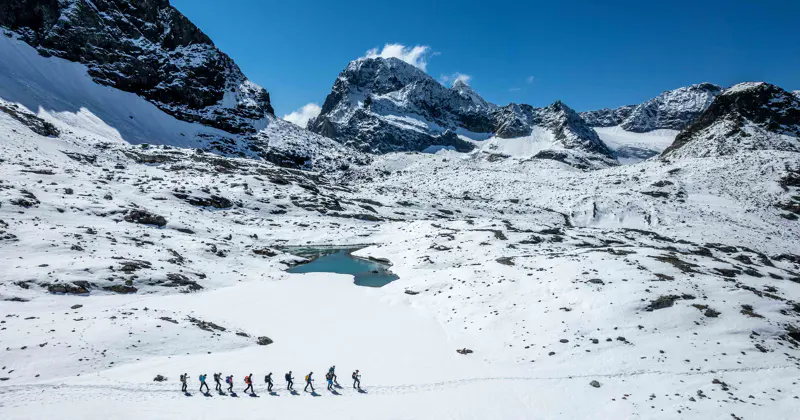 Een groep wandelaars loopt in een rij door een besneeuwd berglandschap met ruige bergtoppen en een klein, gedeeltelijk bevroren meer, onder een helderblauwe hemel.
