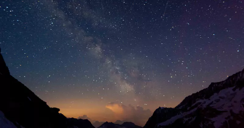 Een sterrenhemel met de Melkweg zichtbaar boven de silhouetten van de bergen; aan de horizon achter de bergtoppen is een vage oranje gloed te zien.