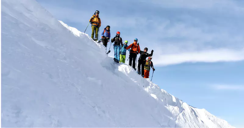 Een groep klimmers in kleurrijke uitrusting staat bij elkaar op een besneeuwde bergkam onder een blauwe hemel; ze houden hun stokken vast en zien er enthousiast uit, met sneeuw en bergtoppen op de achtergrond.