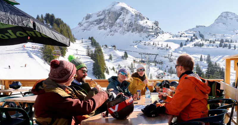 Vijf mensen in winterkleding zitten aan een houten tafel op een besneeuwd bergterras, waar ze eten en praten. Op de heldere, zonnige achtergrond zijn met sneeuw bedekte bergtoppen en skiliften te zien.