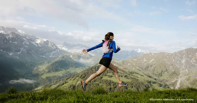 Een vrouw in sportkleding rent over een met gras begroeide heuvelrug, met op de achtergrond besneeuwde bergen en groene valleien onder een gedeeltelijk bewolkte hemel.