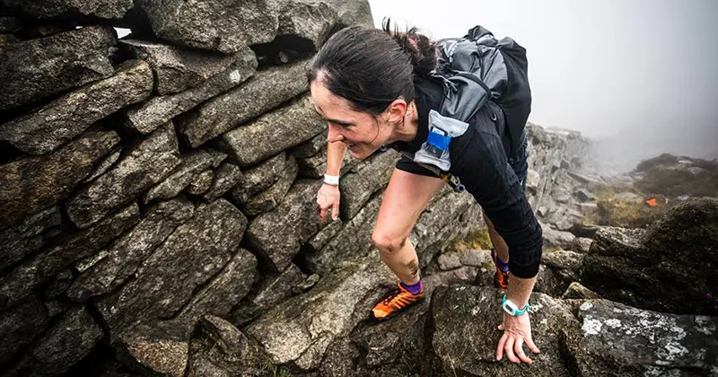 Een vrouw in sportkleding klautert over rotsachtig terrein naast een stenen muur, in mistige, ruige omstandigheden in de buitenlucht.