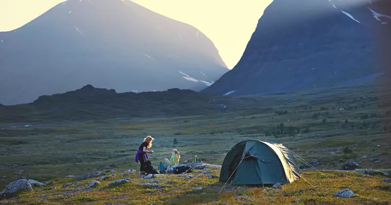 Bij zonsondergang slaan twee mensen hun kamp op met een groene tent in een met gras begroeide vallei, omringd door hoge bergen. In de buurt ligt kampeerspullen verspreid, en het zonlicht schijnt over de bergtoppen op de achtergrond.