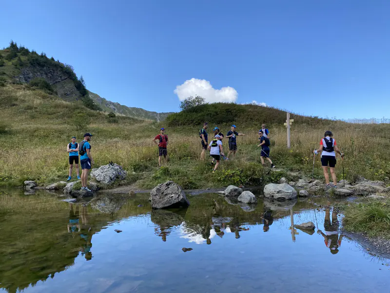 Een groep wandelaars staat te praten bij een klein rotsachtig meertje, met op de achtergrond met gras begroeide heuvels en een blauwe lucht.