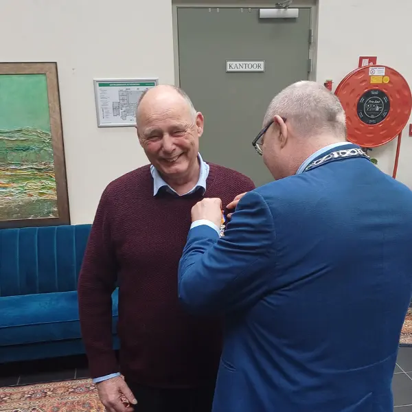 Een glimlachende man krijgt een speldje of medaille van een andere man in een blauw pak. Ze bevinden zich binnenshuis, vlakbij een blauwe bank, een groen schilderij en een rode brandslang. Op een bordje op de deur achter hen staat ‘Kantoor’.
