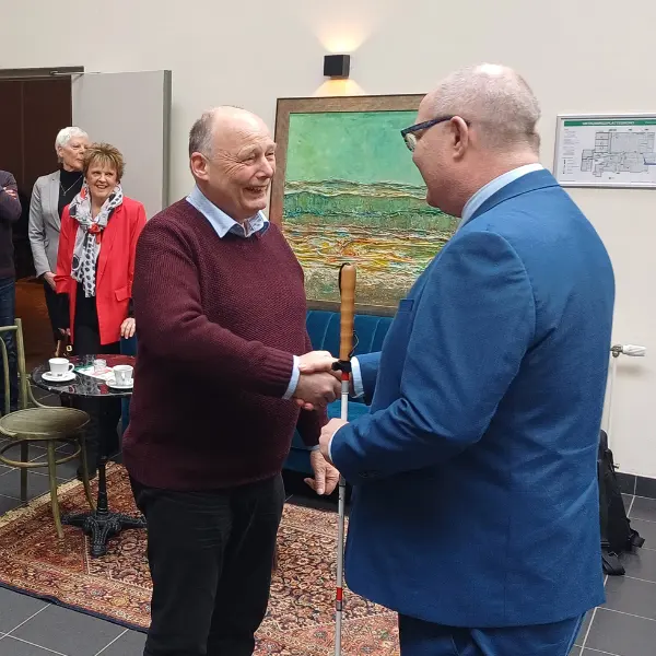 Twee mannen schudden elkaar de hand in een kamer met mensen op de achtergrond. Eén van de mannen houdt een witte stok vast, wat erop wijst dat hij slechtziend is. Aan de muur hangt een schilderij en een kaart, en op de tafels staan koffiekopjes.