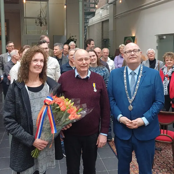 Een vrouw houdt bloemen vast en staat naast twee mannen; de ene draagt een medaille en de andere een blauw pak met een ketting. Achter hen staat een groep mensen in een lichte binnenruimte.