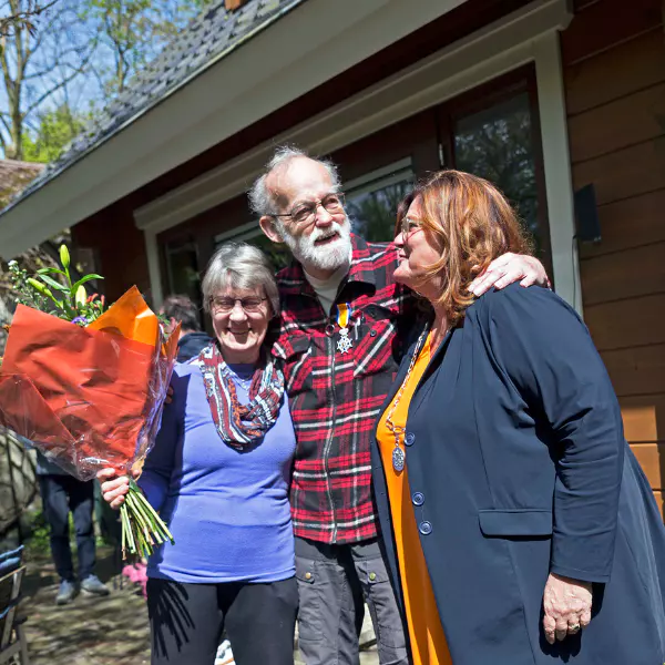 Drie volwassenen staan samen voor een huis. De man in het midden heeft zijn armen om de twee vrouwen heen geslagen. Eén van de vrouwen houdt een groot boeket bloemen vast, en alle drie glimlachen ze; ze zien er blij en feestelijk uit.