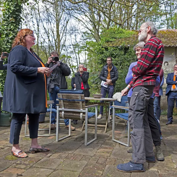 Een vrouw staat buiten tegenover een oudere man, terwijl een aantal mensen toekijkt en een fotograaf foto’s maakt. Ze worden omringd door bomen, lege stoelen en een stenen terras.