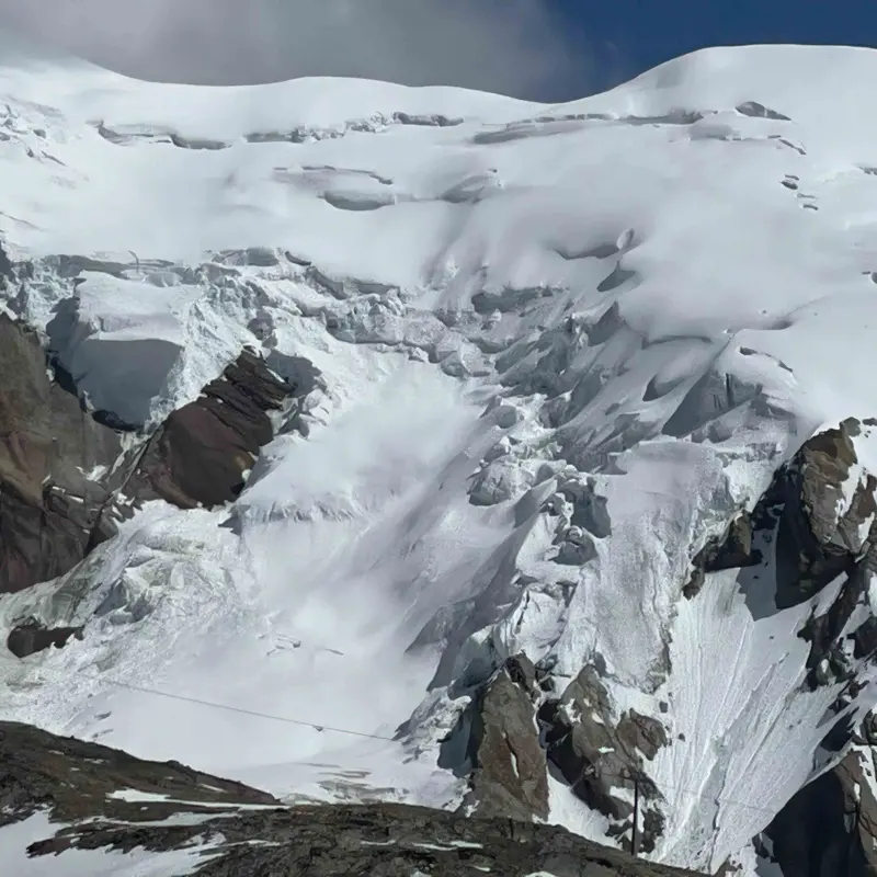 Een met sneeuw bedekte bergtop met ruige hellingen en zichtbare gletsjerspleten, stukken blootliggende rotsen en diepe scheuren. De lucht erboven is gedeeltelijk bewolkt en werpt een zacht licht op het bergachtige terrein.