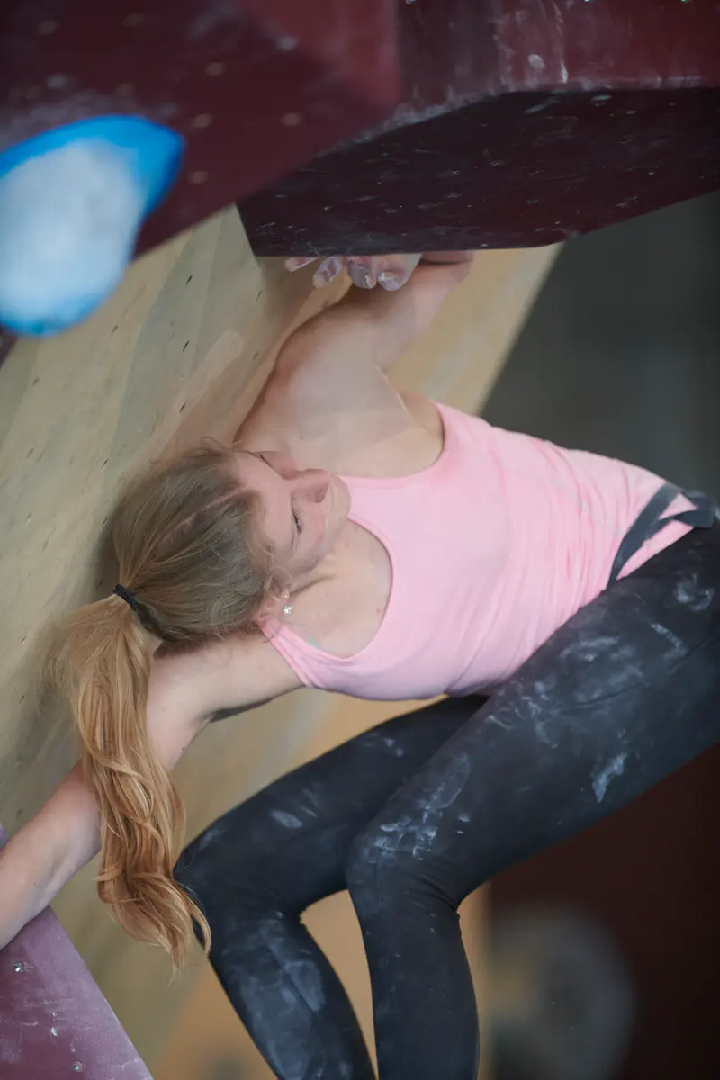 Een vrouw met lang blond haar beklimt een indoor-bouldermuur; ze draagt een roze hemdje en een zwarte broek met krijtvlekken. Ze reikt omhoog en grijpt grepen vast op een steil, schuin houten oppervlak.
