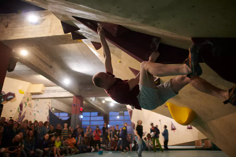 Een klimmer hangt met één hand aan een bouldermuur in een indoorhal, met gestrekte benen, terwijl beneden in de helder verlichte sportzaal een menigte toekijkt.