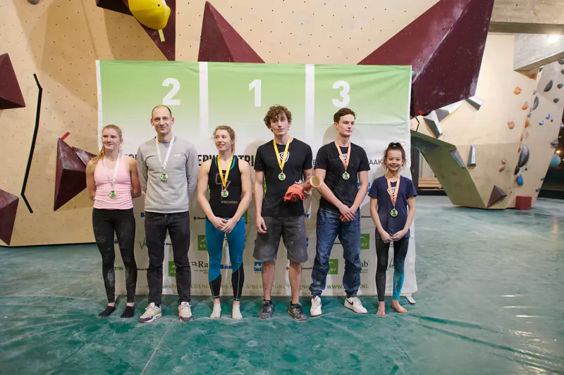 Zes klimmers staan naast elkaar op de vloer van een indoor klimhal, voor een podium met op de achtergrond de nummers 1, 2 en 3. Ze dragen allemaal medailles en sportkleding en poseren voor een groepsfoto na een wedstrijd.