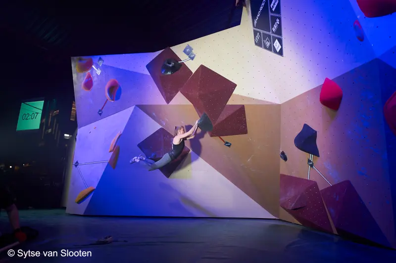 Een klimmer hangt horizontaal aan een kleurrijke bouldermuur in een indoorhal en houdt zich met beide handen vast aan de grepen. Dramatische belichting benadrukt de geometrische panelen van de muur. Op een digitale timer staat 02:07.