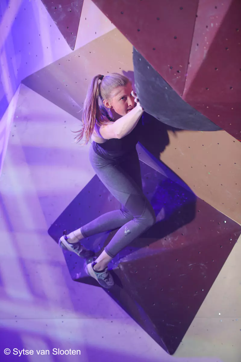 Een vrouw in sportkleding beklimt een indoor-bouldermuur met geometrische grepen, verlicht door paarse lampen.
