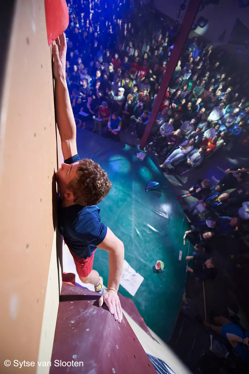 Een klimmer reikt tijdens een wedstrijd naar een greep op een indoor-klimwand, terwijl een groot publiek beneden in een helder verlichte arena toekijkt.