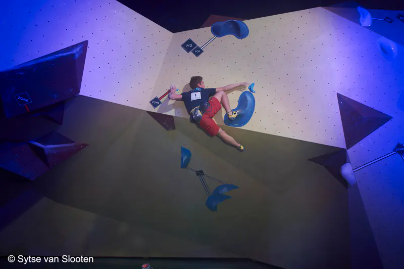 Een klimmer in een rode korte broek beklimt een indoor-bouldermuur; onder dramatische verlichting reikt hij met één hand en één voet naar een greep, terwijl hij zich vasthoudt aan blauwe klimgrepen.