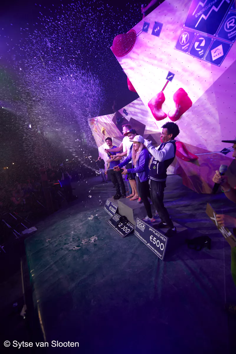 Klimmers vieren na afloop van een wedstrijd hun overwinning op het podium en spuiten champagne in de lucht. Confetti dwarrelt door de lucht, terwijl op de muur achter hen klimgrepen en sponsorlogo’s te zien zijn. Op borden staan de prijsbedragen vermeld.