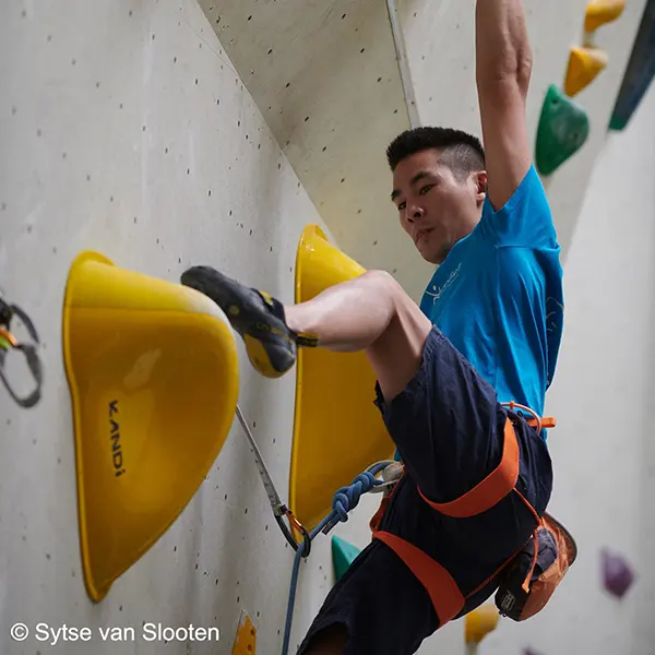 Een klimmer in een blauw shirt en een oranje klimgordel beklimt een indoor-klimwand, waarbij hij zich met zijn handen en voeten vastklampt aan grote gele grepen.