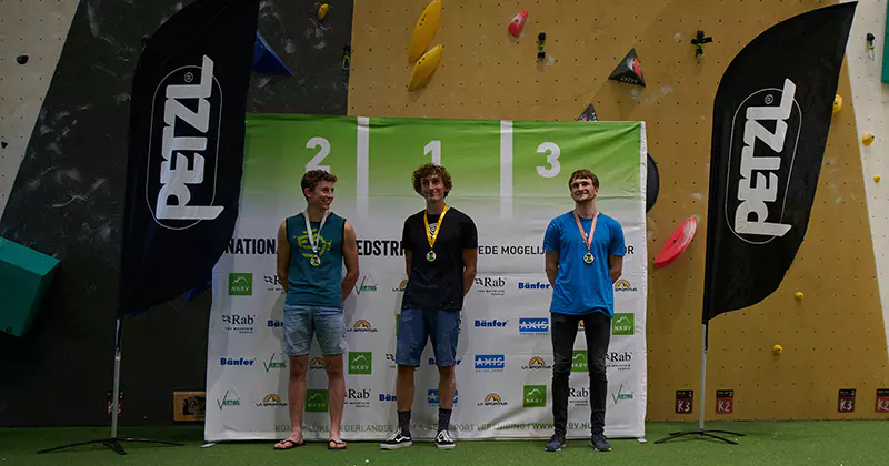 Drie jonge mannelijke klimmers met medailles staan op een podium met de nummers 1, 2 en 3 erop, voor een klimwand en sponsorbanners tijdens een indoor klimwedstrijd.