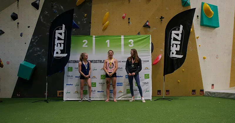 Drie vrouwelijke atleten staan op een podium met bordjes waarop de 1e, 2e en 3e plaats staan aangegeven. Twee vrouwen staan aan de zijkant. Op de achtergrond zijn klimgrepen en vlaggen met merklogo’s te zien.