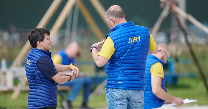 Twee mensen die blauwe hesjes dragen met het opschrift JURY staan buiten met elkaar te praten, terwijl op de wazige achtergrond nog twee anderen zitten. Achter hen zijn een speeltoestel en groen gras te zien.